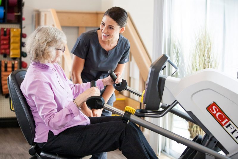 Woman using the scifit machine with the help of a nurse at an assisted living facility 
