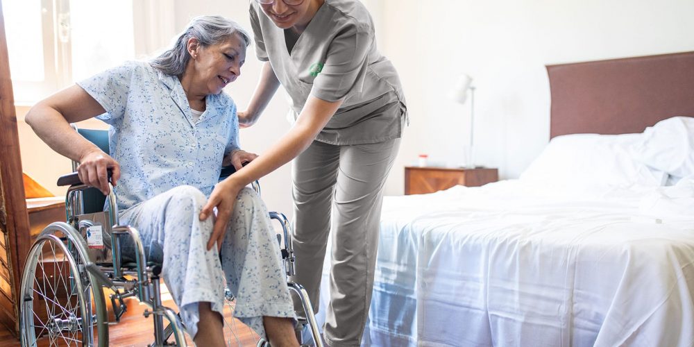 Registered Nurse Helping Elder Woman In Bed From Their Wheelchair