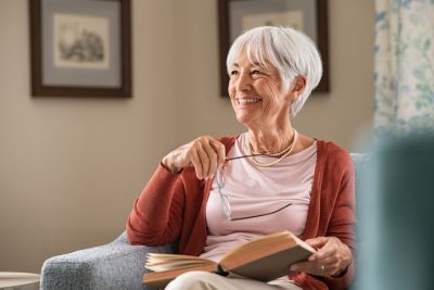 Older woman looking up from book and smiling while holding glasses in one hand