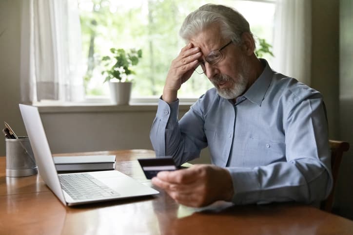 Older man sitting near laptop looking at credit card shocked 