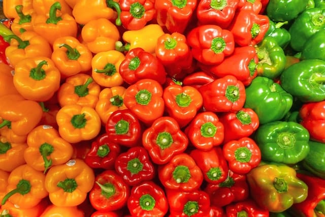 An image filled with colored peppers. From left to right: orange peppers, red peppers, green peppers.