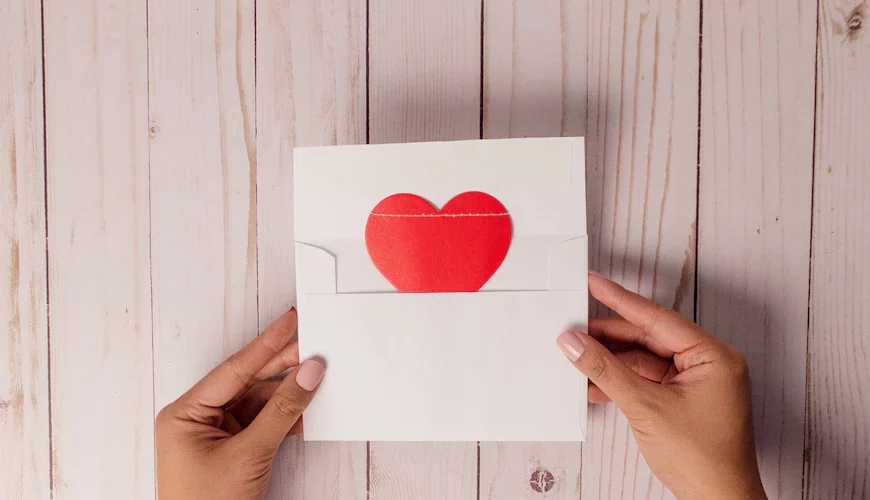 Two hands holding a white envelope with a white card peeking out of the top that contains a red heart in the center.