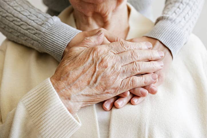 Focus is on the hands of an elderly person, clasping their hand over an individual who is giving them a hug from behind.