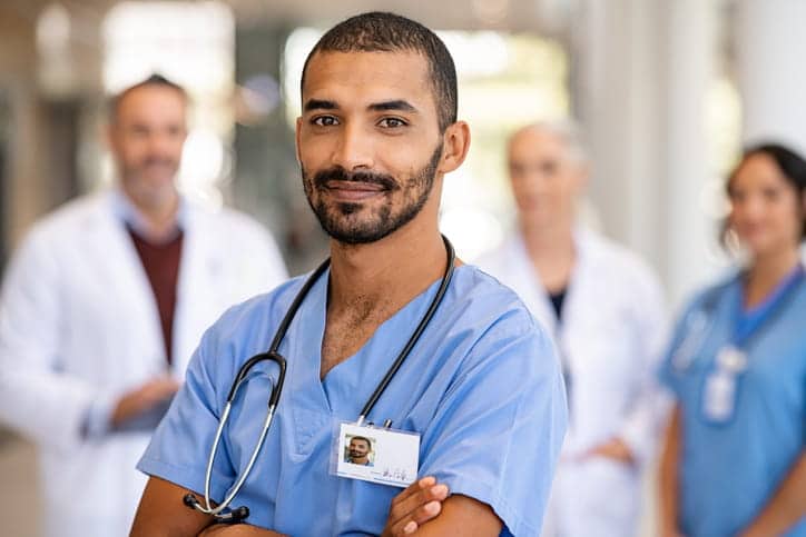 A male CNA with his arms crossed, smiling, after finishing CNA training. Other medical personnel are in the background, blurred out.