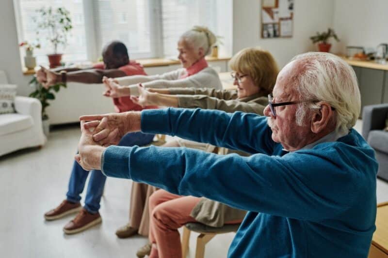 Four Seniors Doing Stretching Exercises With Their Arms While Sitting Down.