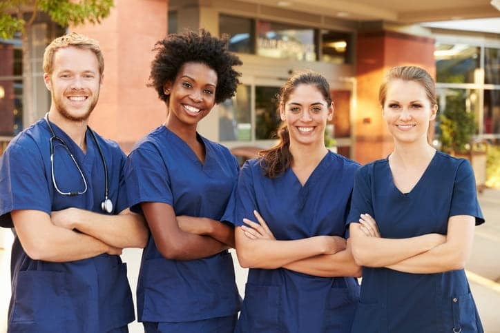 Four nurses in navy scrubs posing in a row with their arms crossed in front of a skilled nursing facility.