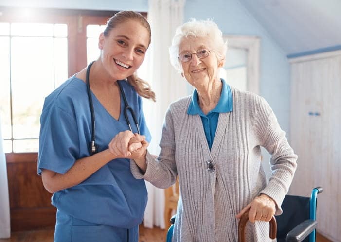 A nurse holds the hand of an elderly woman at a skilled nursing facility. They are smiling together for a photo.