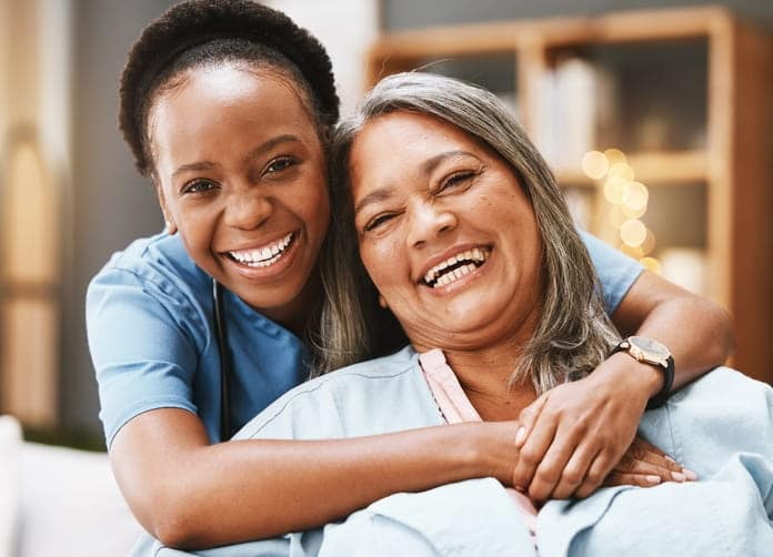 A nurse smiling with her arms around an elderly patient as she gives her a hug from behind at a skilled nursing facility.