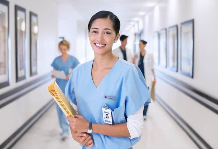 A nurse smiling at a skilled nursing facility while holding patient charts. In the background are three employees blurred out.
