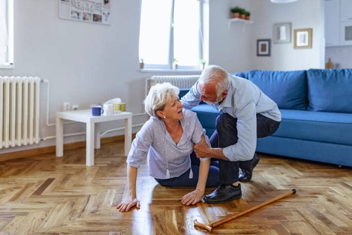 An elderly male helping his wife up off the ground after she fell in their living room. 