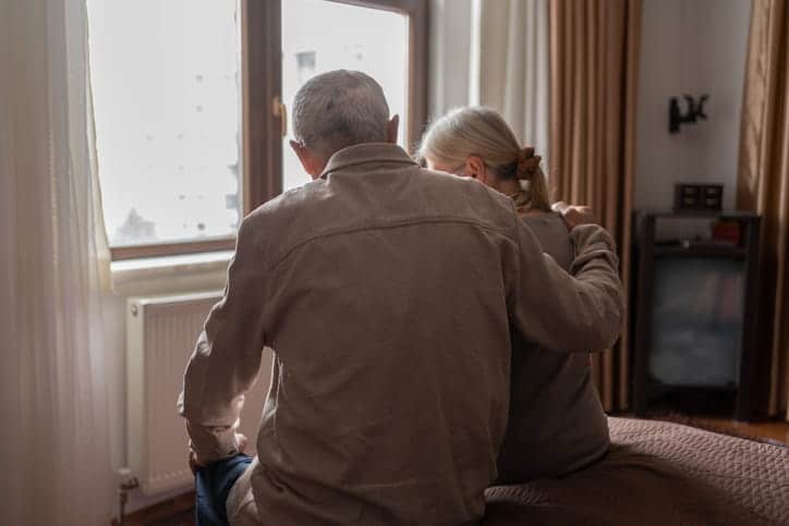 The backside of a man wrapping an arm around his wife as they sit on the edge of the bed. He is taking care of her as she suffers from dementia.
