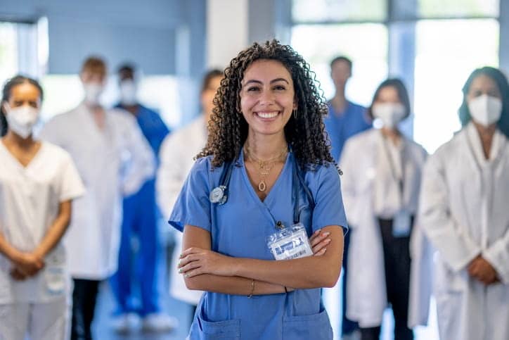 A nurse smiling with her arms crossed. Behind her, out of focus are more nurses and doctors at a skilled nursing facility. 