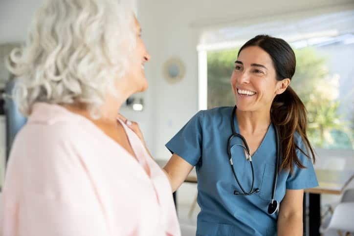 An aid is smiling and speaking with an elderly patient at a skilled nursing facility. 