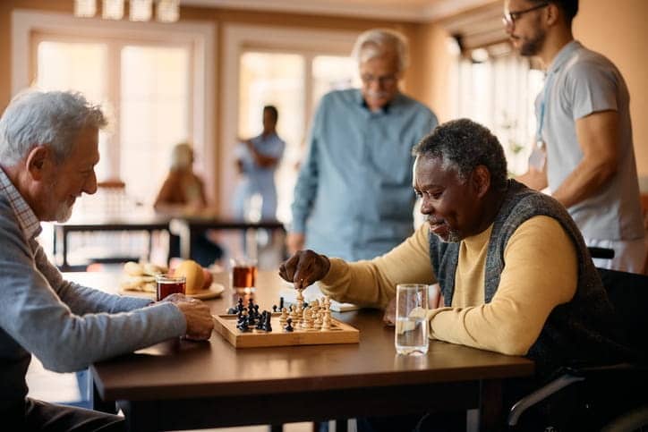 Two elderly men playing chess at an assisted living facility. In the background is an aid helping another patient. 