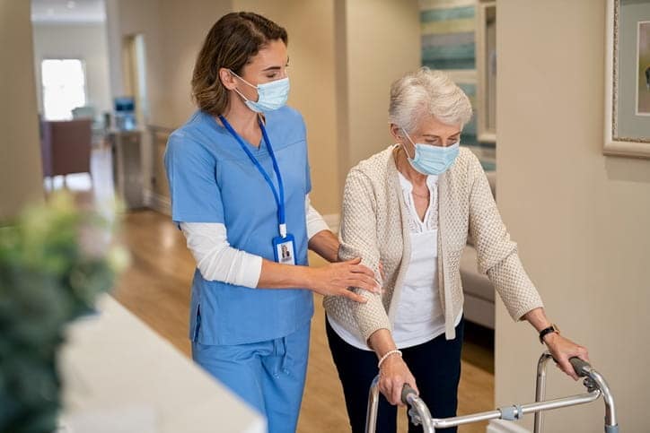 A nurse walking with an elderly patient with a walker in a skilled nursing facility. They are both wearing face masks.