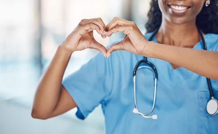 A nurse wearing blue scrubs is smiling and making a heart with her hands. 