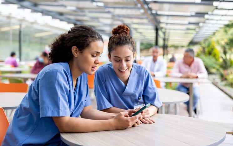 Two nurses at a table looking at a cellphone. 