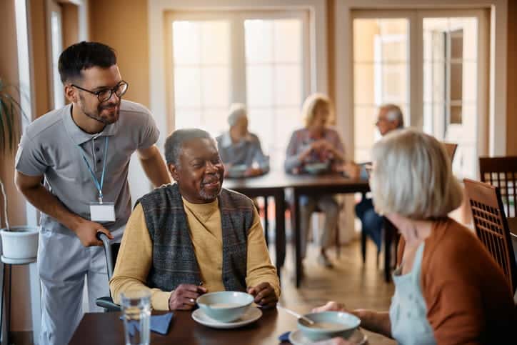 Two patients enjoying lunch together in a skilled nursing facility as a worker joins them. 