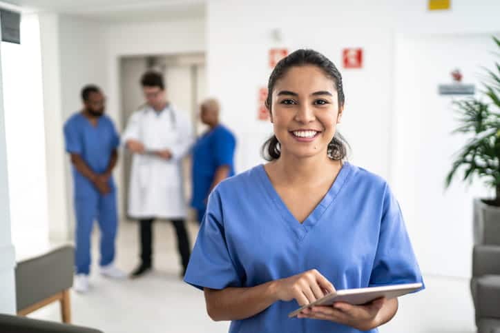 A nurse holding a tablet, smiling for the camera. In the background are three more workers blurred out. 