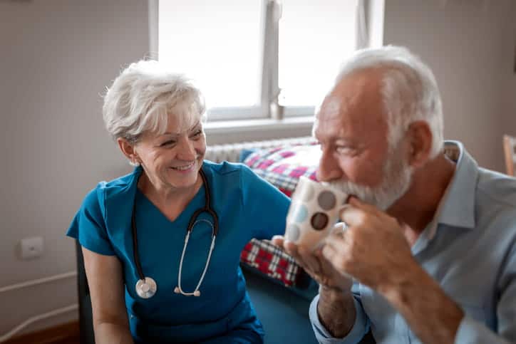 A nurse sitting with an elderly man as he drinks from a coffee mug. 