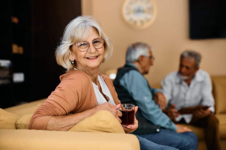 A woman smiling in assisted living with two other patients talking in the background. 