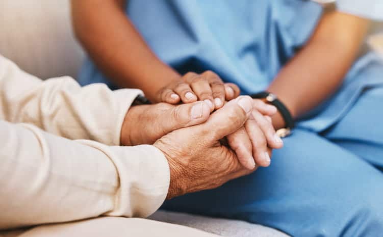 An assisted living facility aid holding hands with an elderly man as she comforts him. 