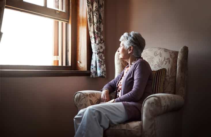 An elderly lady sits in a chair as she gazes out the window.