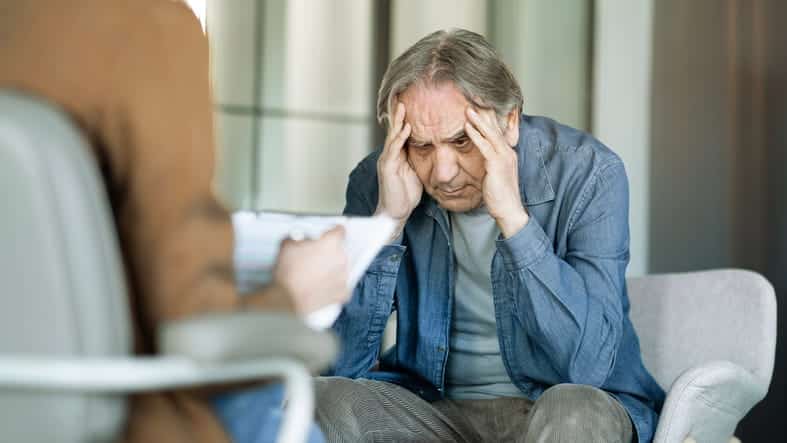 An elderly man holding his head in frustration as he speaks with a counselor. 