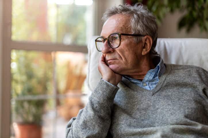 An elderly man rests his head in his hand as he looks out the window.