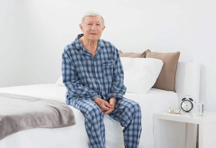 An elderly man wearing pajamas as he sits on the edge of his bed. 