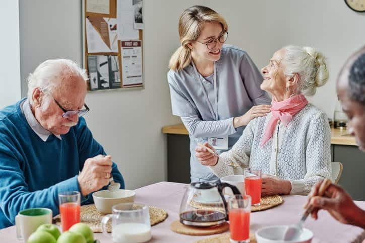 nurse talking to older woman in wheelchair at dining table with other residents of assisted living facility