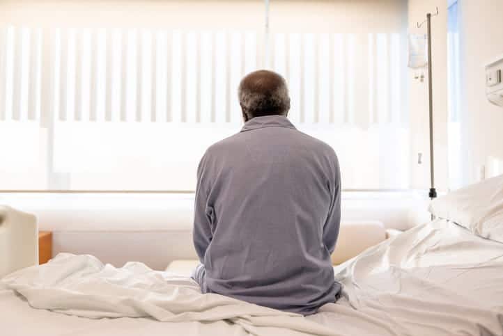 The back of an elderly man sitting on the edge of his bed.