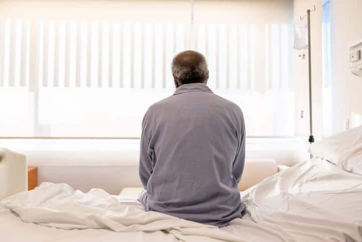 The back of an elderly man sitting on the edge of his bed. 