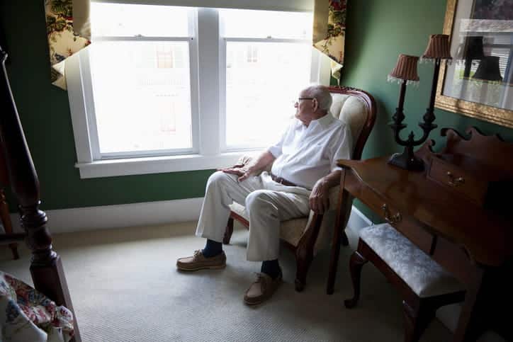 An elderly man sits in a chair alone while looking out a window.
