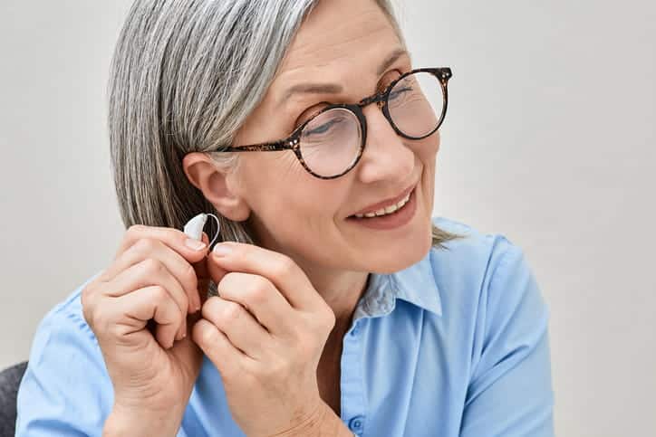An elderly woman with glasses putting in her hearing aid.
