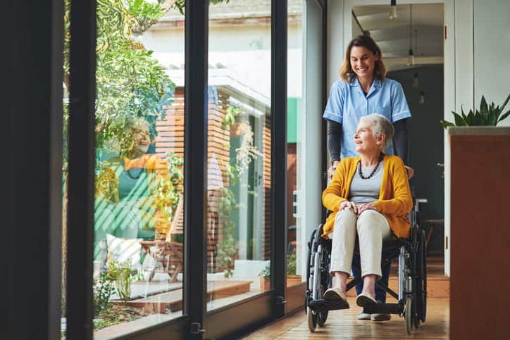An Assisted Living Worker Pushing An Elderly Woman In A Wheelchair Through The Facility.