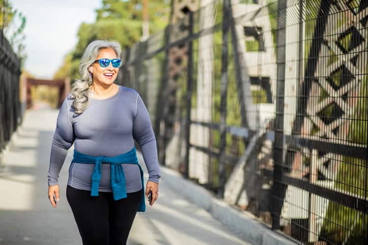 A Woman Smiling As She Walks In Seasonal Weather. She Has A Long Sleeve With A Hoodie Tied Around Her Waist And Is Also Wearing Sunglasses. 