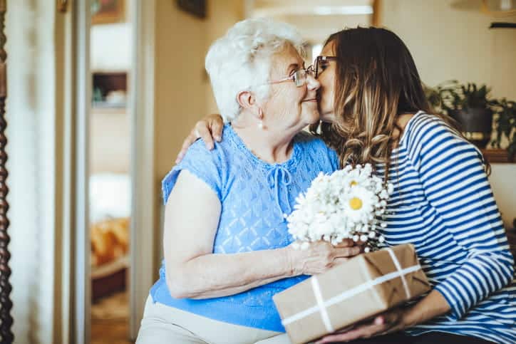 A Daughter Hugging Their Elderly Mother And Kissing Her On The Cheek As She Gives Her A Present. 