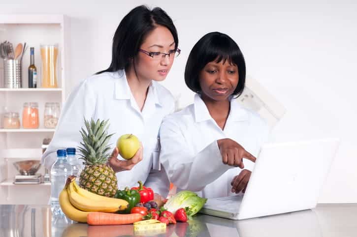 Two Dietitians Looking At A Computer Screen Next To A Pile Of Fruits To Make A Diet Plan For A Client. 