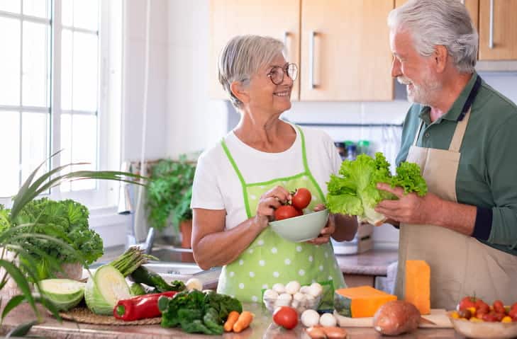 An Elderly Couple In Their Kitchen Surrounded By Fruits And Vegetables To Eat Healthier. 