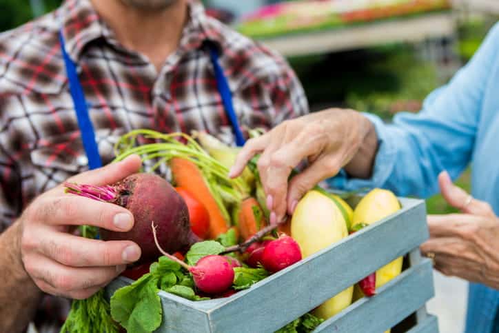 A Man Holding A Fresh Crate Of Vegetables At A Farmers' Market As An Elderly Woman Inspects It. 
