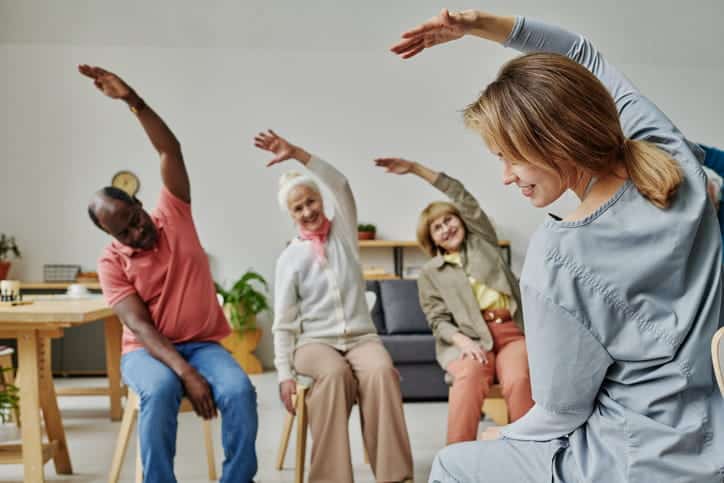 A Health Aide Is Leading A Stretching Class For Three Seniors In An Assisted Living Facility. 
