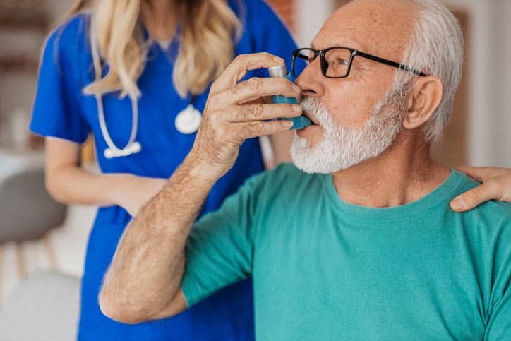 A Nurse Helping An Elderly Man As He Uses His Inhaler Due To Respiratory Issues.