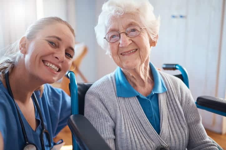 A Nurse Smiling With An Elderly Female In A Wheelchair At A Nursing Home.
