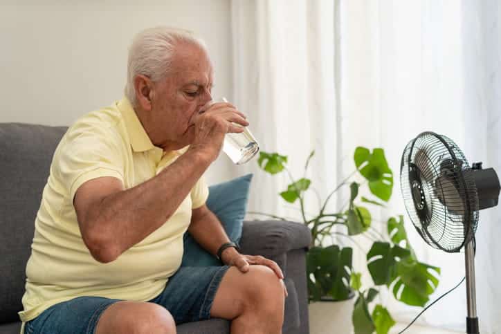 A Senior Drinking A Glass Of Water While Sitting In Front Of A Fan To Help Cool Down From Heat Exposure.