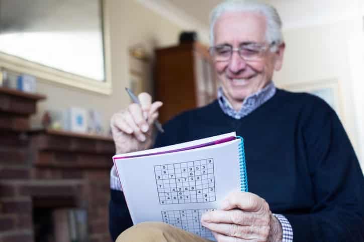 A Senior Smiling As He Works On A Sudoku Puzzle.