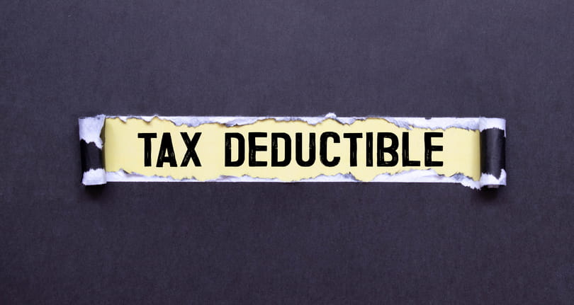 The Words &Quot;Tax Deductible&Quot; In Black Capital Letters On A Yellow Background, Revealed Through A Horizontal Tear In A Sheet Of Dark Gray Paper.