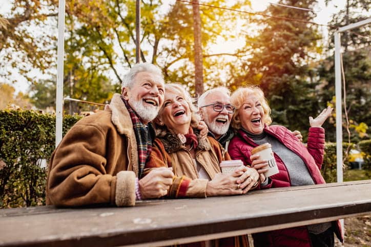 A Joyful Group Of Four Senior Friends—Two Men And Two Women—Are Laughing Heartily While Gathered At A Wooden Table Outdoors On A Sunny Autumn Day.