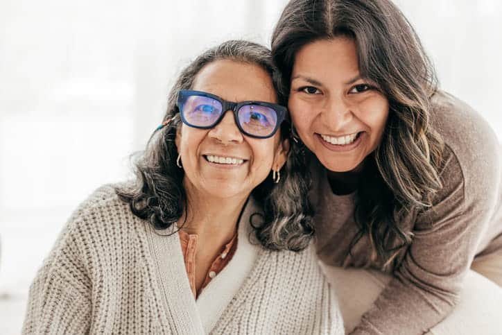A Warm Portrait Of A Senior Hispanic Woman Wearing Glasses And Her Adult Daughter Smiling Together. The Daughter Is Affectionately Leaning In From Behind Her Mother, And Both Are Looking At The Camera.