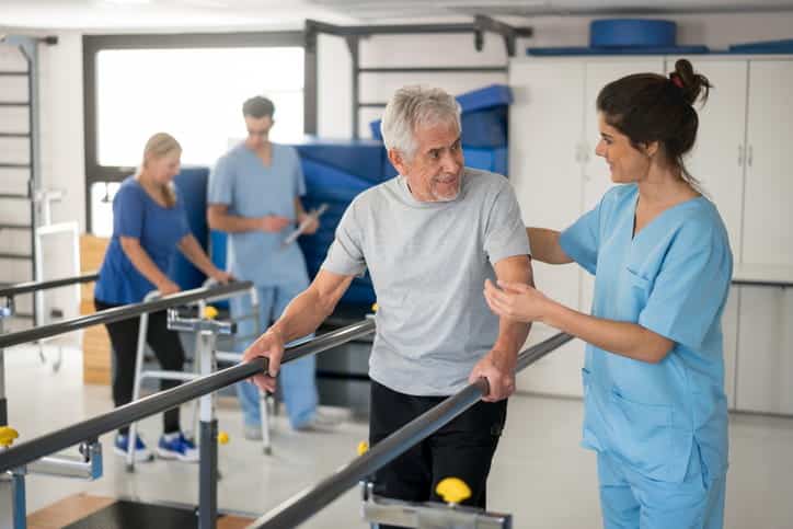 A Female Physical Therapist In Scrubs Assists A Senior Man As He Uses Parallel Bars For Walking And Balance Exercises In A Rehabilitation Clinic.